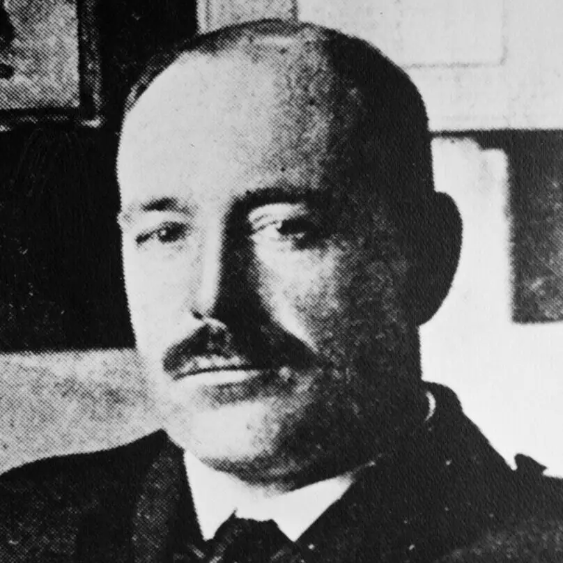A historic black‑and‑white portrait of a mustached man in a suit and tie, photographed indoors with framed items behind him. The image captures early‑20th‑century style, formal attire, and classic portrait lighting, making it ideal for heritage, history, or archival content.